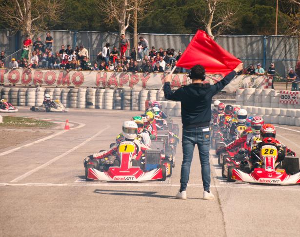 Gara di kart con piloti pronti alla partenza e bandiera rossa alzata.